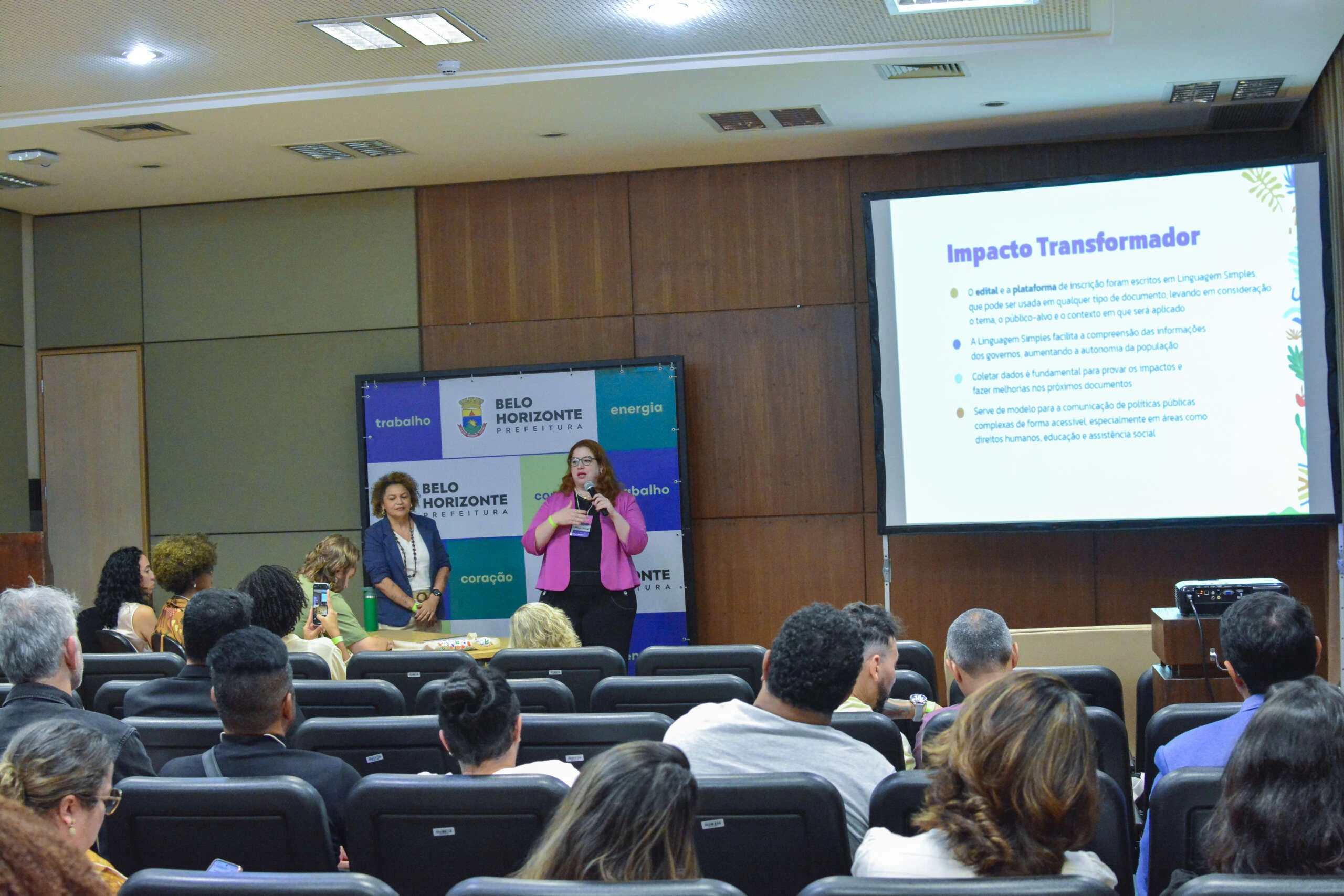 Apresentação em auditório durante o 5º Prêmio Conexão Inova, promovido pela Prefeitura de Belo Horizonte. Duas mulheres estão em pé no palco: uma ao centro com microfone fazendo apresentação, e outra ao lado, ouvindo. Ao fundo, painel com logotipo da Prefeitura e palavras como "trabalho", "energia" e "coração". À direita, telão exibe o slide intitulado "Impacto Transformador", com tópicos sobre linguagem simples, autonomia da população e comunicação acessível. O público assiste atento, sentado em cadeiras pretas.