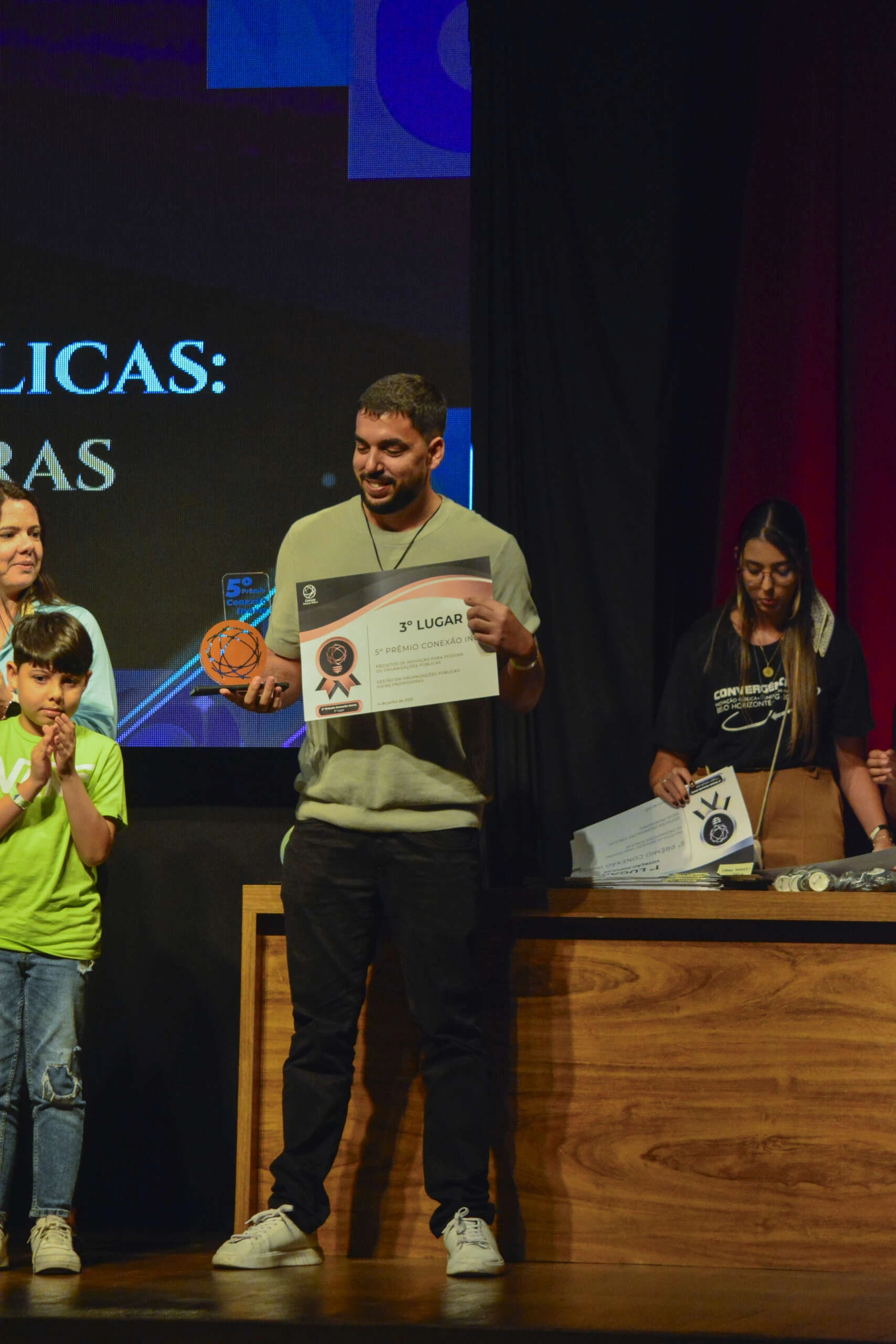 Homem sorri no palco segurando um troféu laranja e um certificado de "3º Lugar" do 5º Prêmio Conexão Inova. À esquerda, uma mulher e uma criança assistem à premiação; à direita, uma mulher atrás de uma bancada organiza documentos com o mesmo selo do prêmio. O evento ocorre em um auditório com iluminação de palco, e o clima é de celebração.