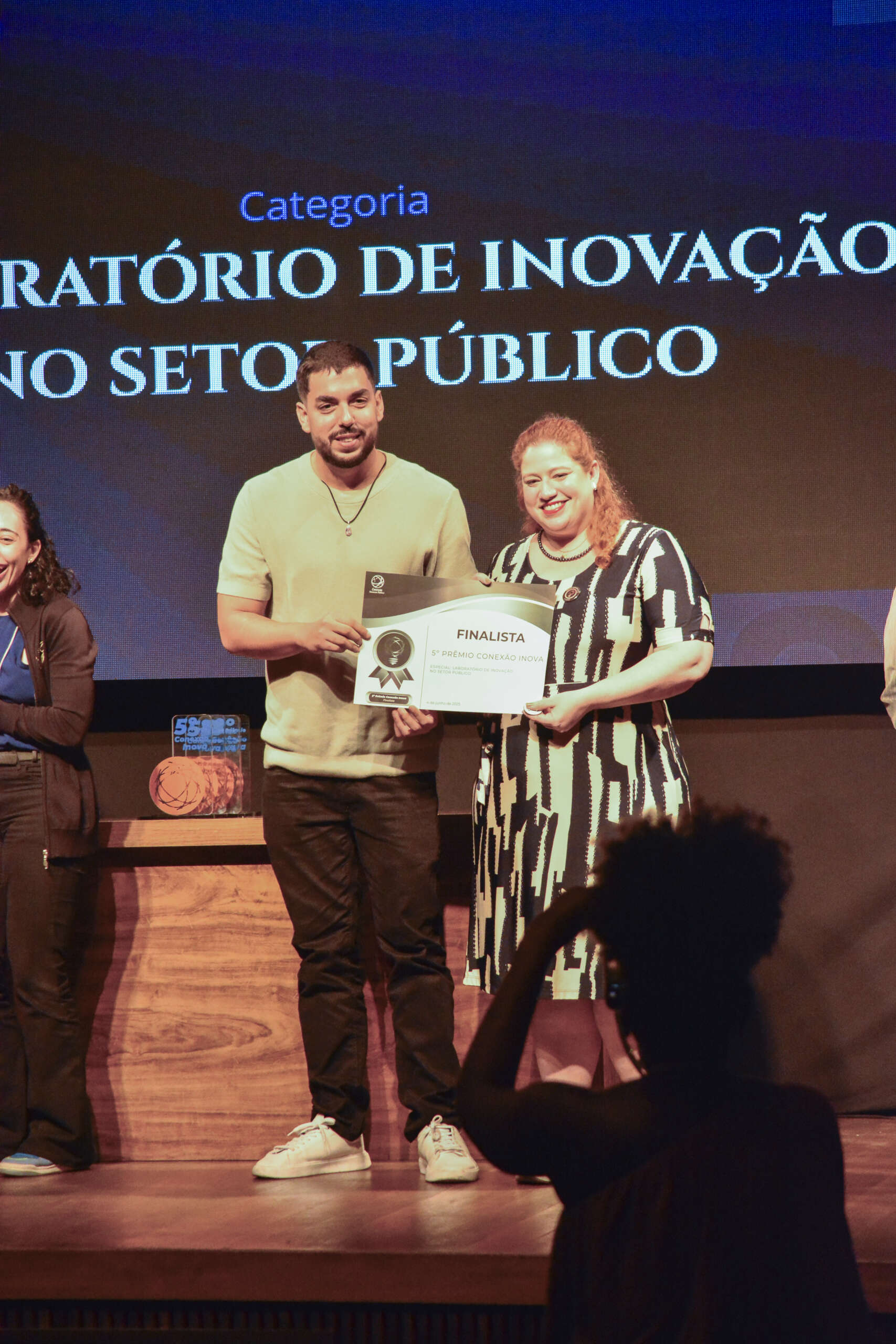 Duas pessoas em pé no palco durante cerimônia do 5º Prêmio Conexão Inova, segurando um certificado com o selo de “Finalista”. Atrás deles, o telão exibe a frase “Categoria: Laboratório de Inovação no Setor Público”. Ambos sorriem enquanto posam para foto. À esquerda, parte de outra participante aparece; à frente do palco, uma pessoa com cabelo preso em coque fotografa a cena. Sobre a bancada, um troféu laranja do prêmio está visível.