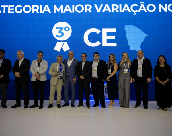 Grupo de 9 pessoas, incluindo homens e mulheres, em um palco. Ao fundo, um telão azul exibe o texto "Maior variação nominal’, 3º lugar, estado do Ceará.” e o mapa do Ceará.