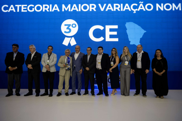 Grupo de 9 pessoas, incluindo homens e mulheres, em um palco. Ao fundo, um telão azul exibe o texto "Maior variação nominal’, 3º lugar, estado do Ceará.” e o mapa do Ceará.