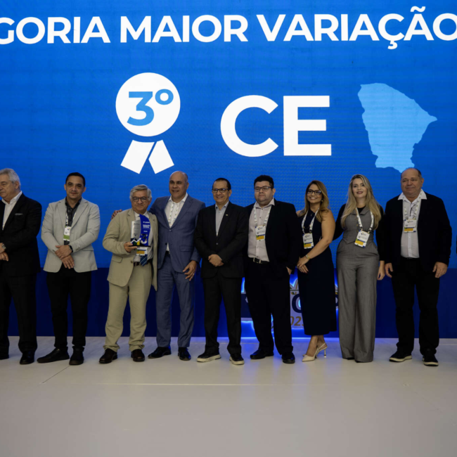 Grupo de 9 pessoas, incluindo homens e mulheres, em um palco. Ao fundo, um telão azul exibe o texto "Maior variação nominal’, 3º lugar, estado do Ceará.” e o mapa do Ceará.