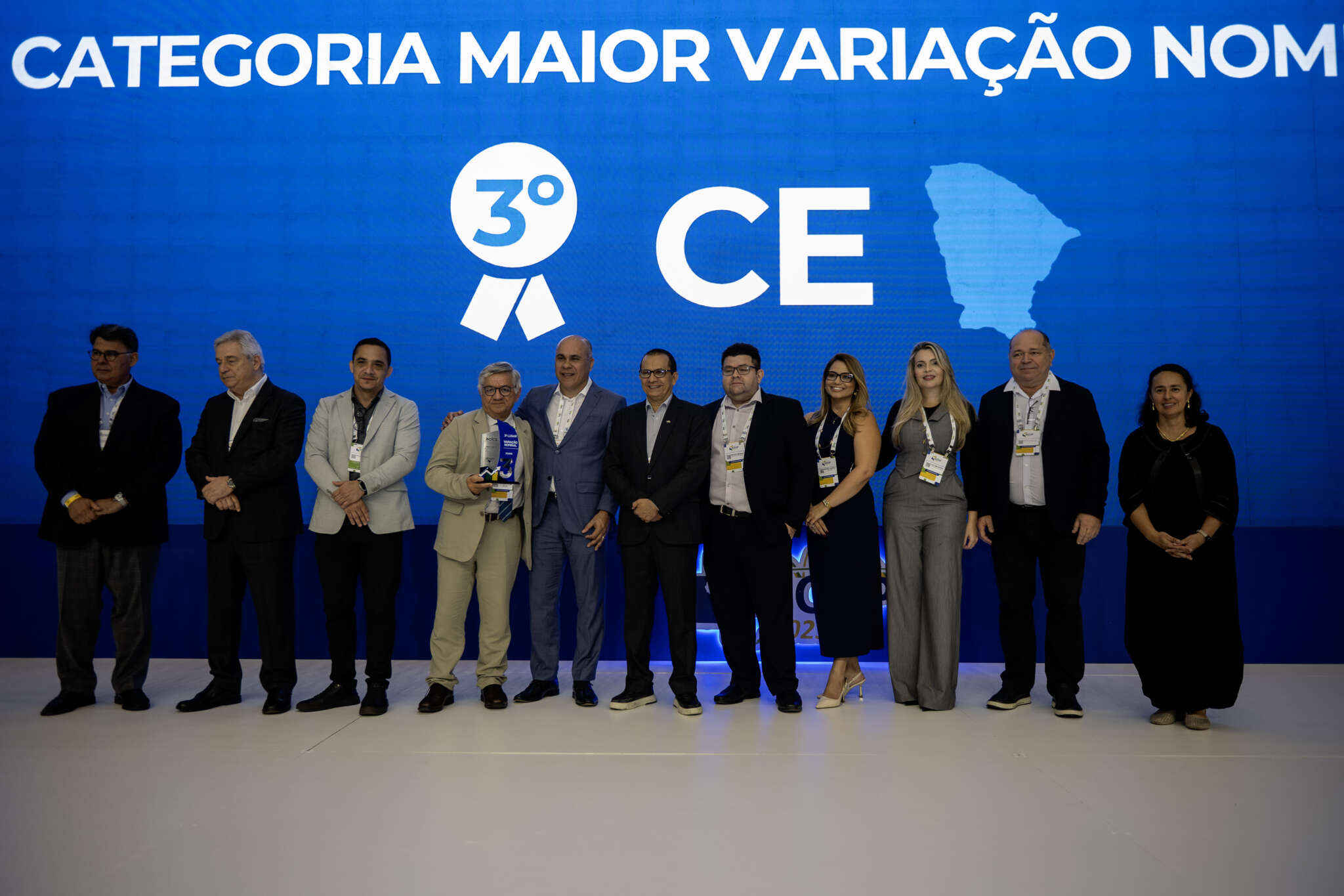 Grupo de 9 pessoas, incluindo homens e mulheres, em um palco. Ao fundo, um telão azul exibe o texto "Maior variação nominal’, 3º lugar, estado do Ceará.” e o mapa do Ceará.