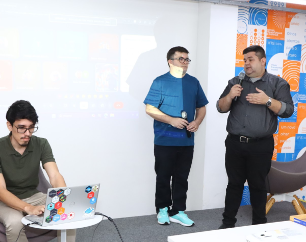 Três homens, dois em pé com microfones e um sentado com notebook, em sala de apresentação.