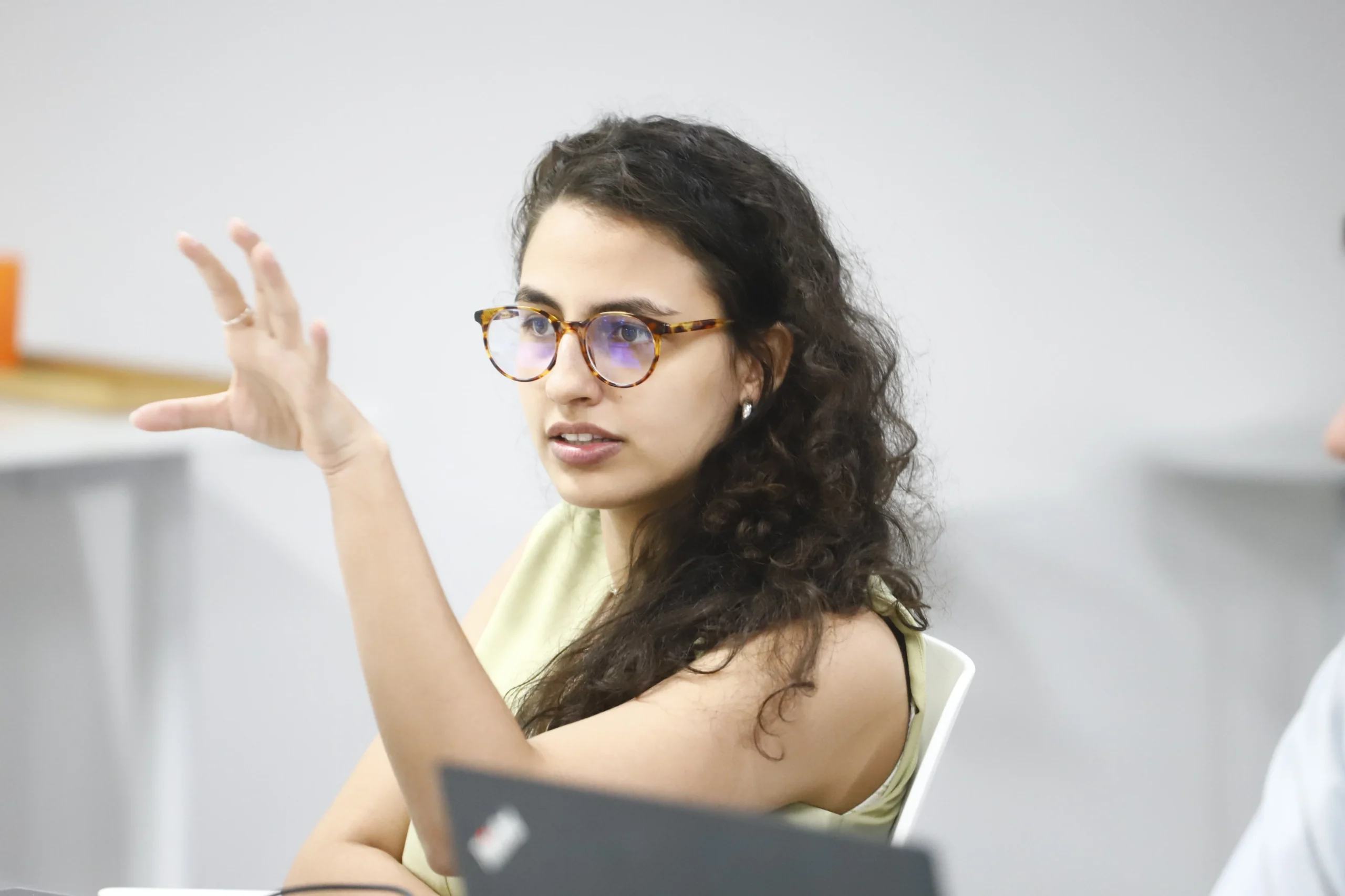 Mulher com cabelos cacheados e óculos, vestindo uma blusa clara. Ela está sentada em uma mesa de reunião e gesticula com a mão esquerda enquanto fala.