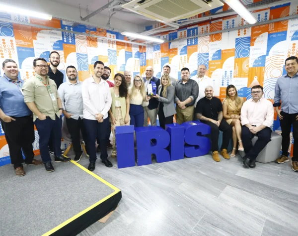 Grupo de cerca de 18 pessoas, incluindo homens e mulheres, de pé e sentadas ao redor de um letreiro azul com a palavra 'IRIS'. O fundo é uma parede com padrão geométrico em tons de azul e laranja com a mesma palavra.