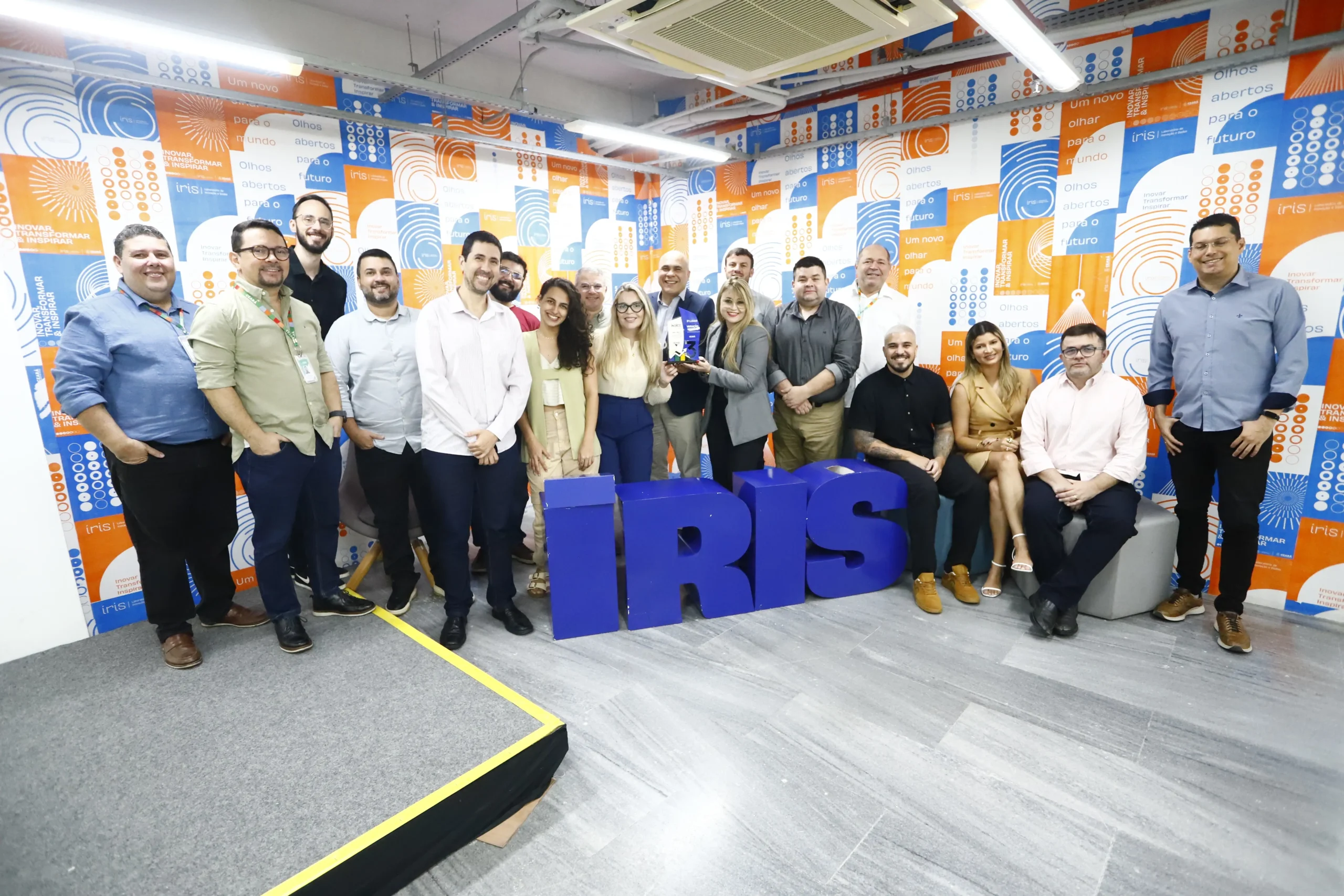 Grupo de cerca de 18 pessoas, incluindo homens e mulheres, de pé e sentadas ao redor de um letreiro azul com a palavra 'IRIS'. O fundo é uma parede com padrão geométrico em tons de azul e laranja com a mesma palavra.