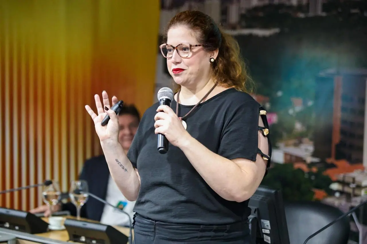 Mulher com óculos e camisa preta segurando um microfone e um controle remoto, gesticulando enquanto fala durante uma apresentação.