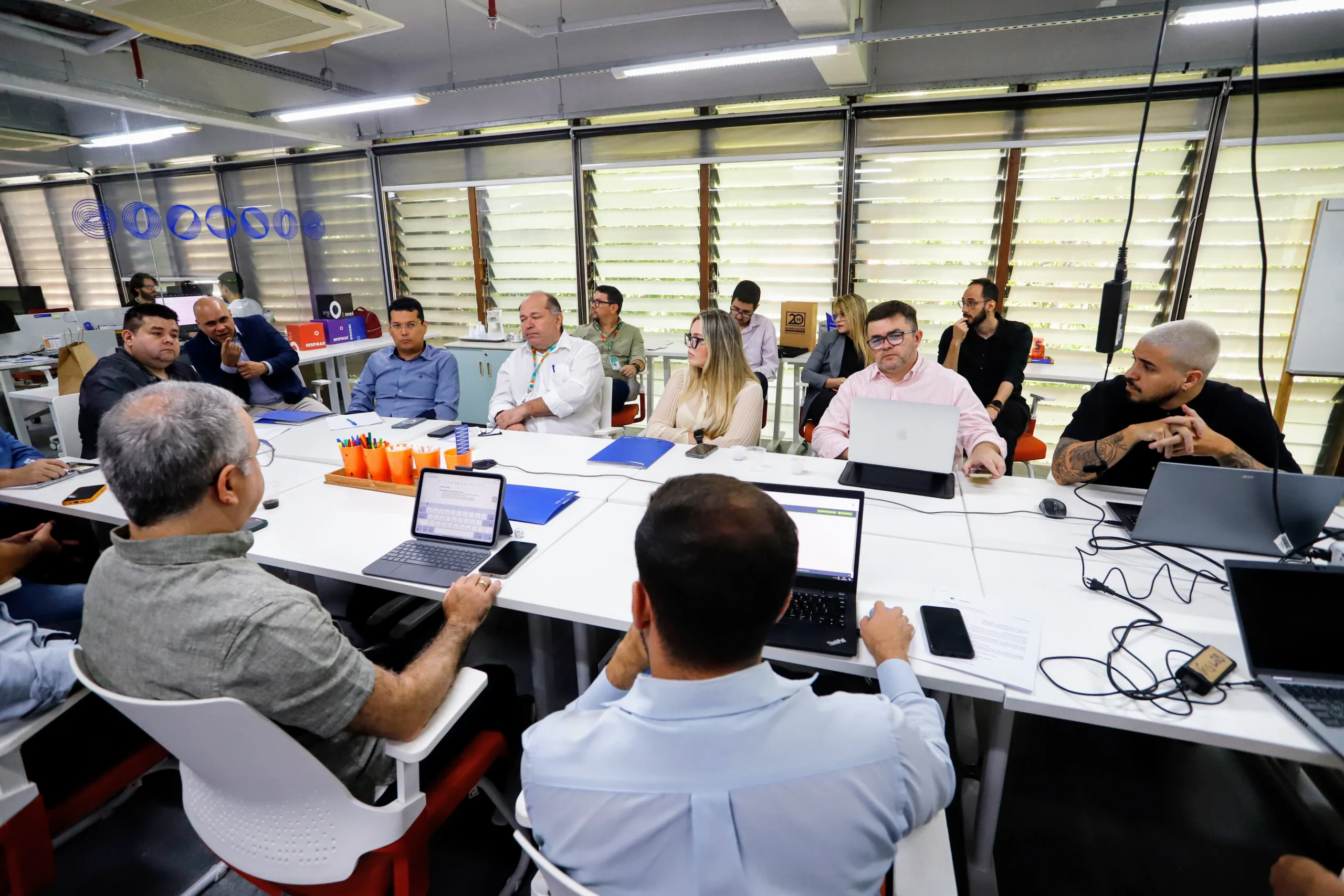Reunião no espaço multimídia. Homens e mulheres estão sentados em uma grande mesa branca. Há notebooks, celulares e copos na mesa.