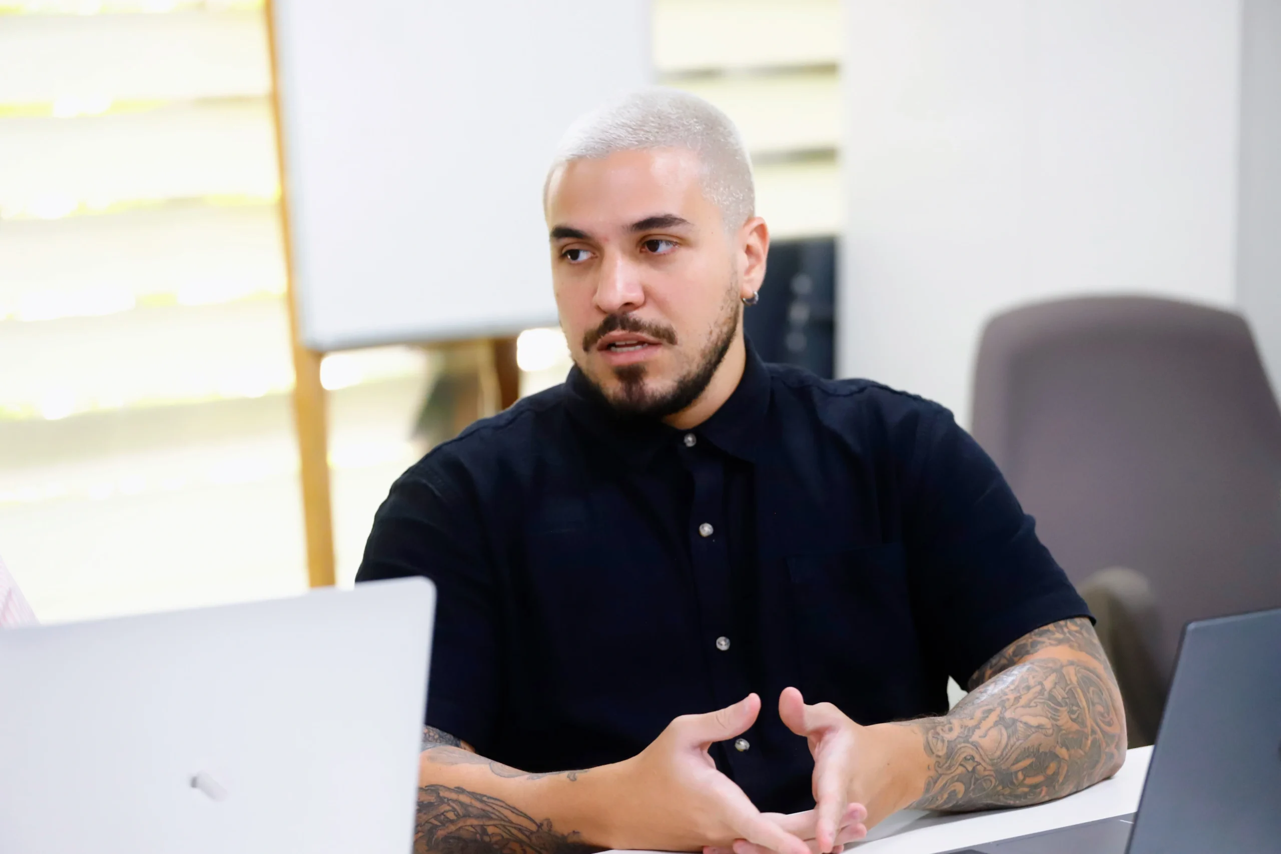 Homem com cabelo loiro e barba, usando uma camisa preta, sentado em uma mesa de reunião e gesticulando com as mãos. Ele tem tatuagens nos braços.