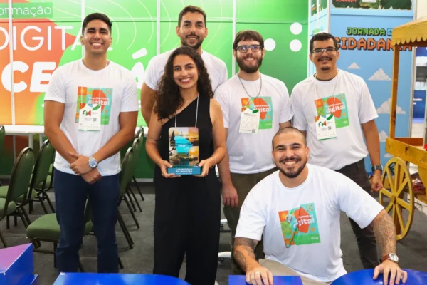 Seis participantes em estande com painéis ‘Transformação Digital no Ceará’ e ‘Jornada da Cidadania’; uma mulher ao centro segura um troféu.