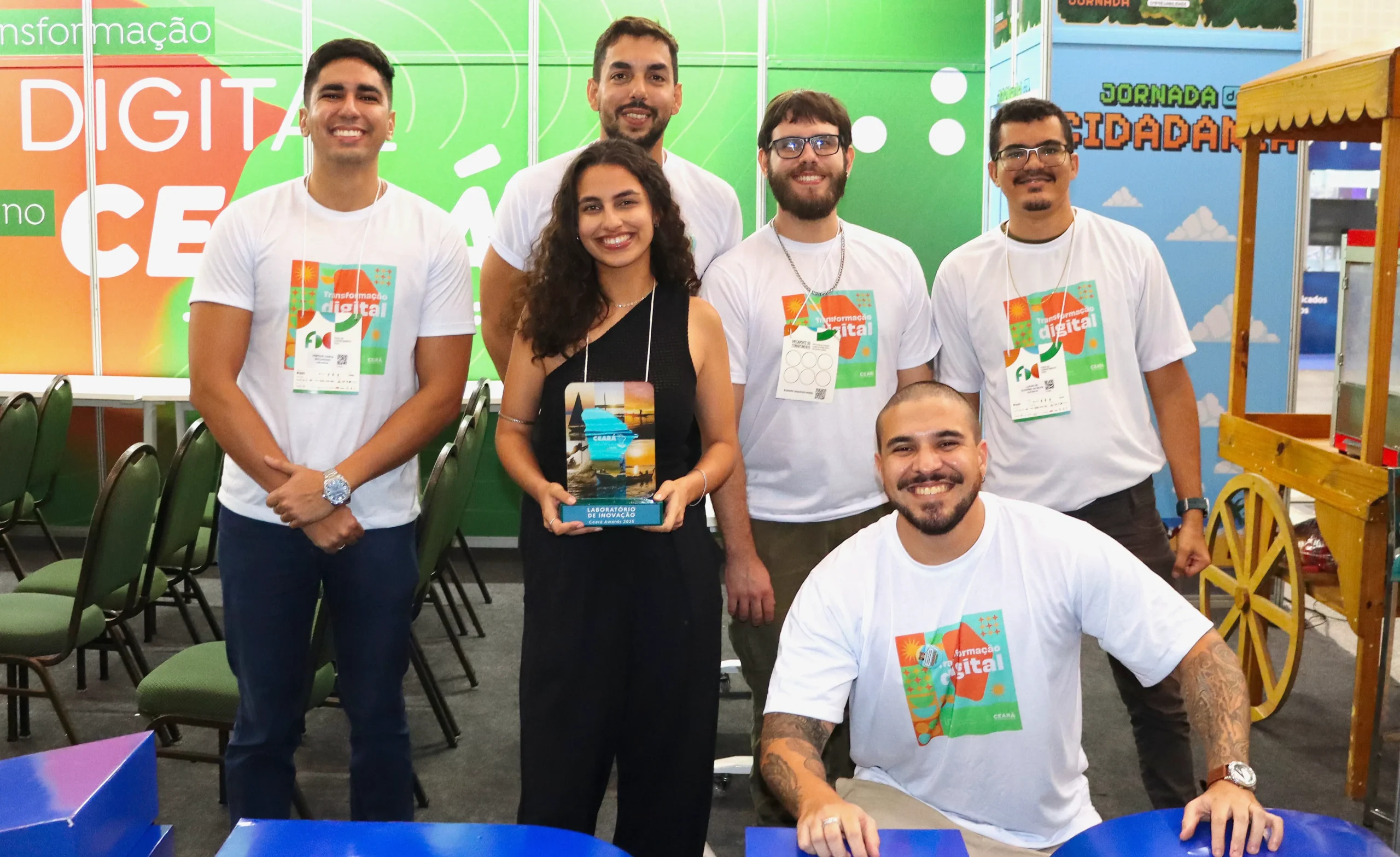 Seis participantes em estande com painéis ‘Transformação Digital no Ceará’ e ‘Jornada da Cidadania’; uma mulher ao centro segura um troféu.