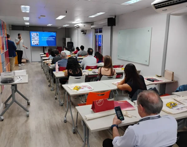 Homem de pé palestrando em sala de treinamento lotada com pessoas sentadas em mesas olhando a tela de projeção.