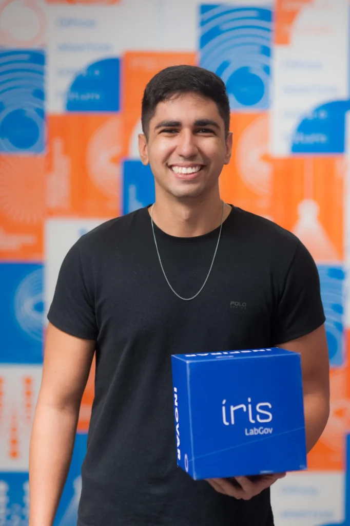 Líder técnico Vinicius sorrindo, usando camiseta preta, em frente a uma parede institucional com padrões circulares em azul e laranja. Ele segura uma caixa azul que exibe o logo do Íris LabGov.