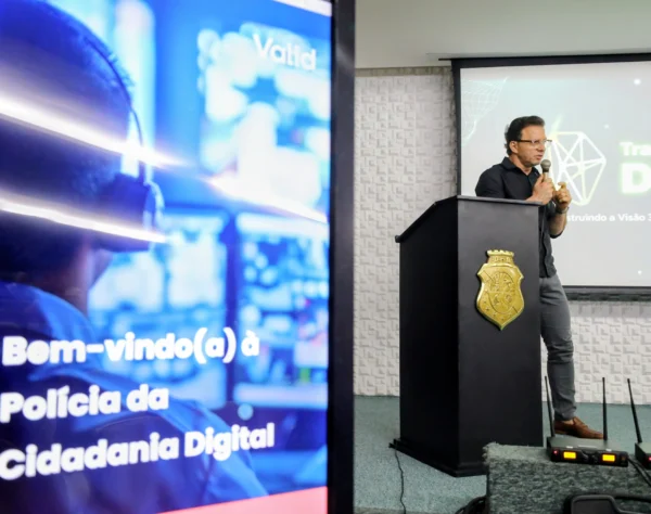 Acessor Vidal, realizando palestra, de camisa preta e óculos, falando ao microfone atrás de um púlpito com o brasão do governo do estado do ceará. O monitor lateral exibe a mensagem 'Bem-vindo(a) à Polícia da Cidadania Digital'.