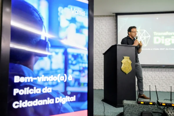 Acessor Vidal, realizando palestra, de camisa preta e óculos, falando ao microfone atrás de um púlpito com o brasão do governo do estado do ceará. O monitor lateral exibe a mensagem 'Bem-vindo(a) à Polícia da Cidadania Digital'.