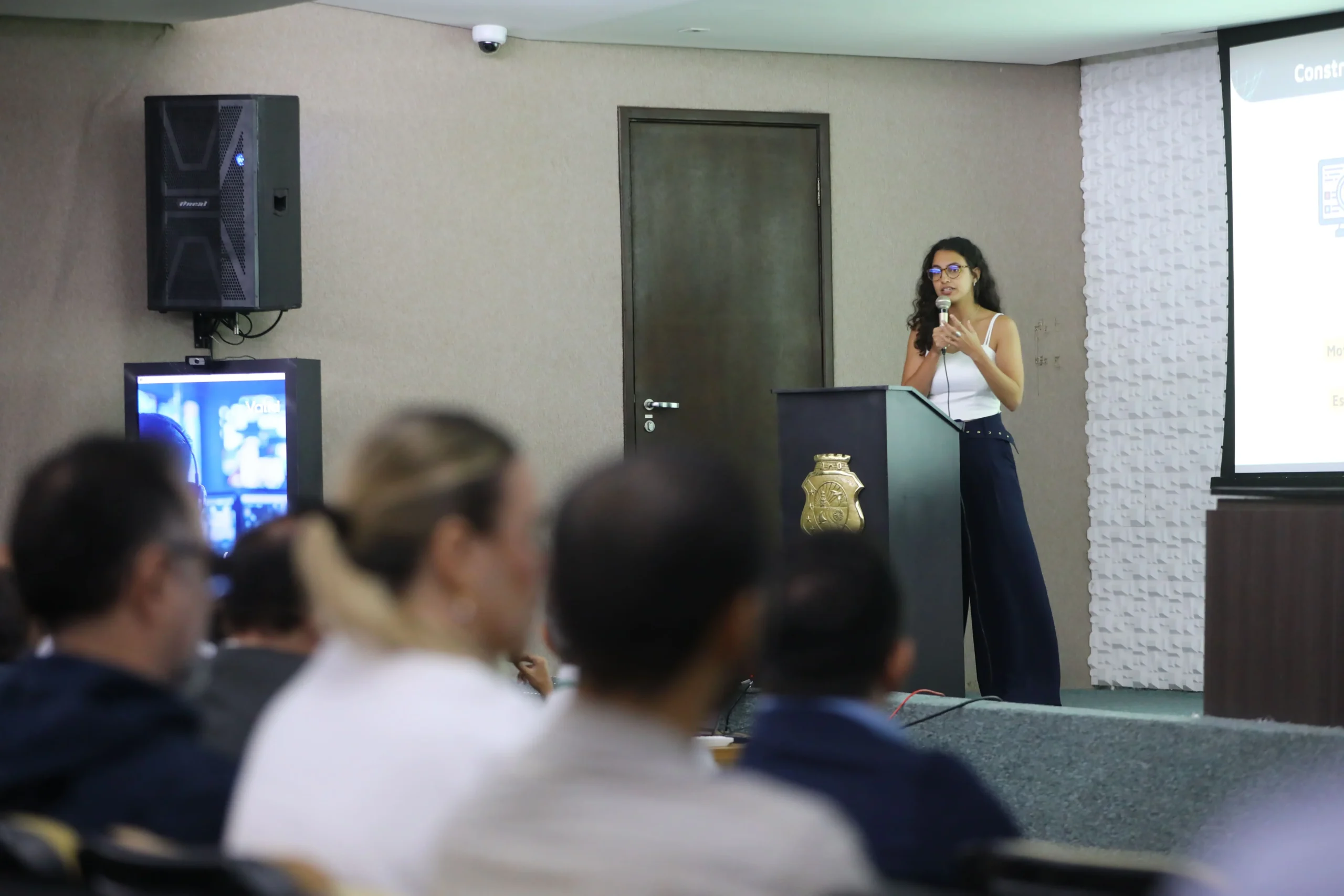 coordenadora geral do íris luiza clara pacheco, vestindo top branco e calça escura de cintura alta, falando ao microfone em um púlpito escuro com brasão dourado do governo do estado do ceará, para uma plateia com foco na nuca dos participantes.